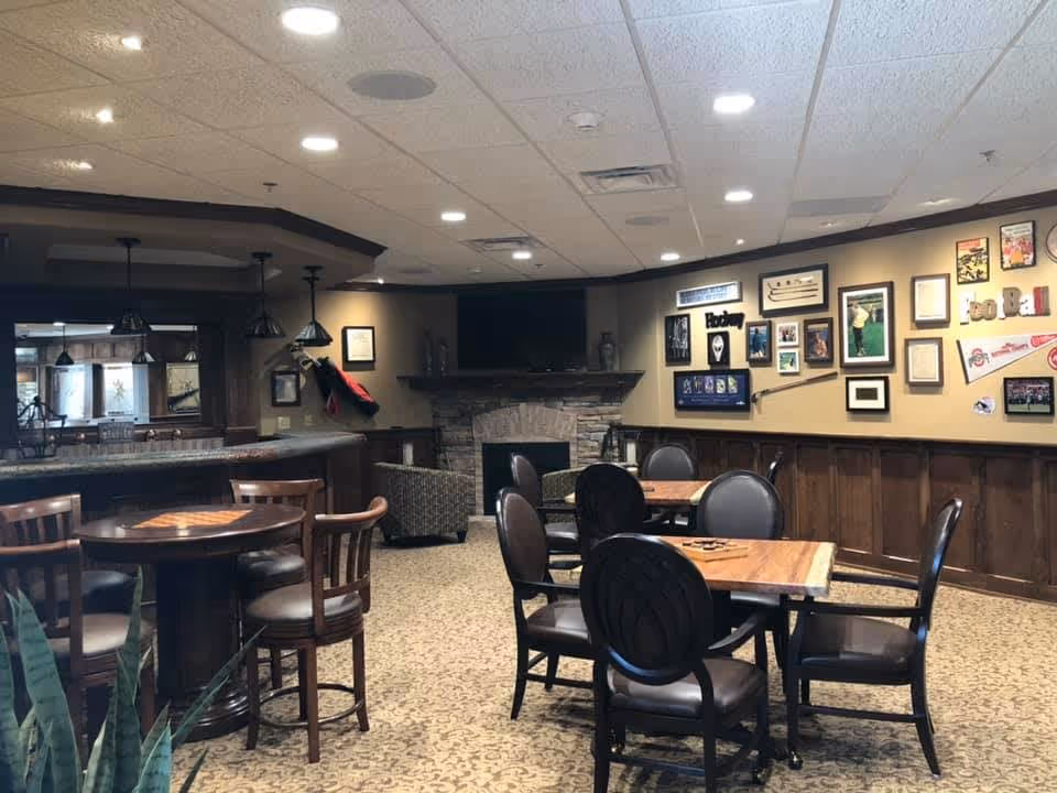 Community lounge with tables and chairs, a stone fireplace and wall-mounted TV, and a bar counter with pendant lights.