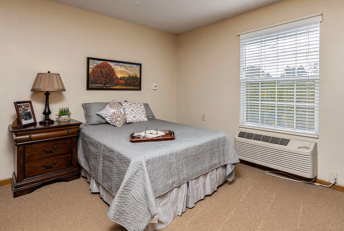 A bedroom with a neatly made bed covered with a gray quilt and three decorative pillows. On the bed is a wooden tray holding an open book and a pair of glasses. To the left of the bed is a wooden nightstand with a table lamp, a framed photo, and a small potted plant. Above the bed hangs a framed picture of a tree with autumn foliage. To the right is a window with white blinds and an air conditioning unit below it. The walls are beige and the floor is carpeted.