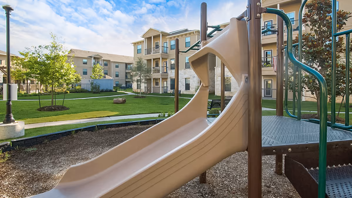 Playground slide and climbing structure in an outdoor courtyard area with green grass, trees, and a multi-story residential building in the background under a partly cloudy sky.