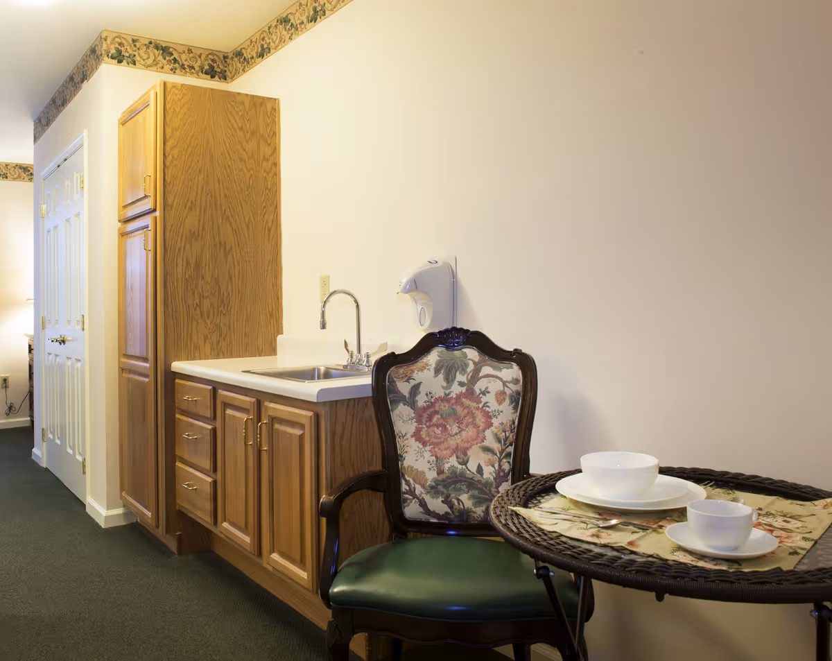 Small kitchenette with wood cabinets and sink beside an upholstered chair and a small round table set with dishes.