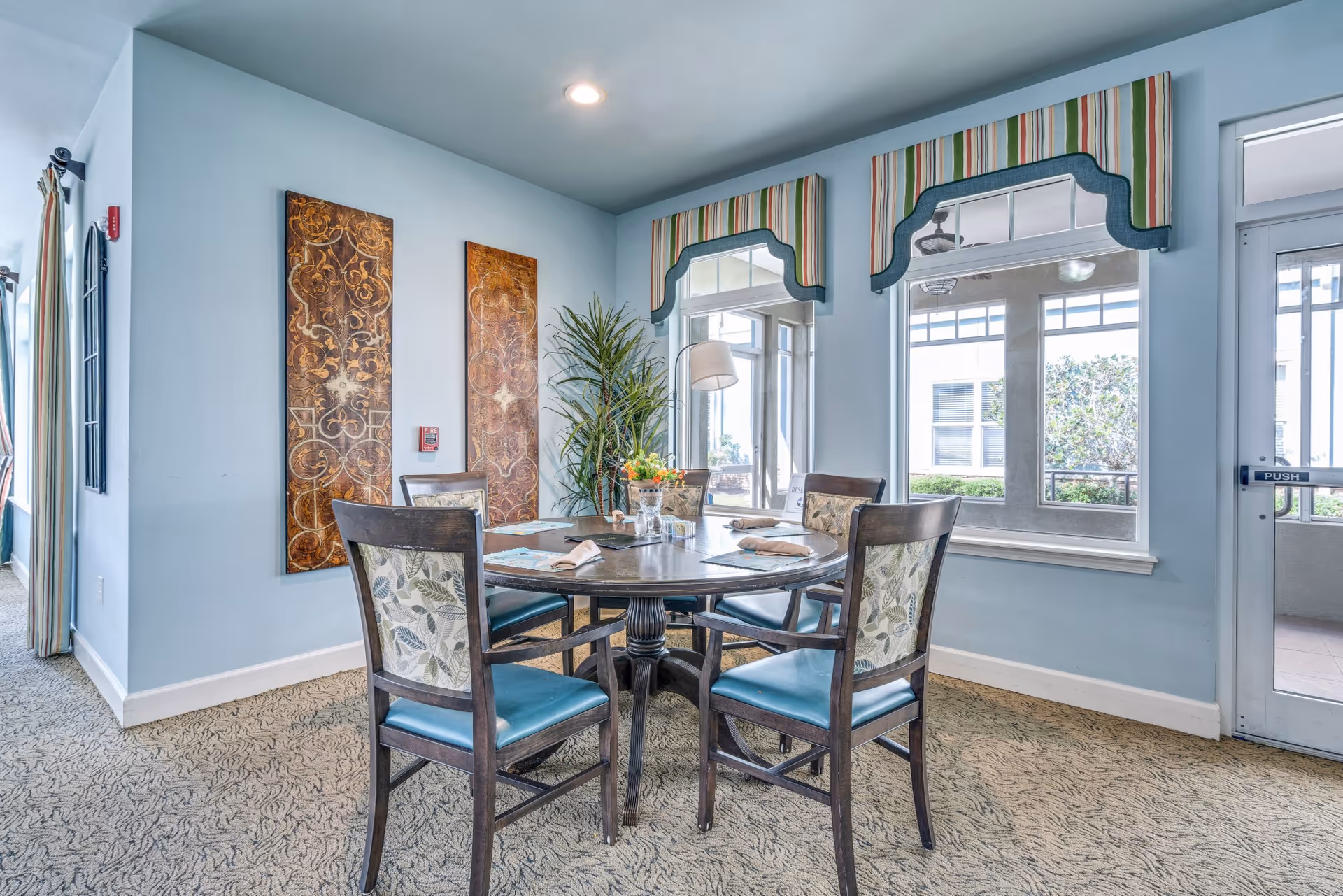 A dining area with a round wooden table set with placemats and napkins, surrounded by four chairs with blue seats and floral-patterned backs. The room has light blue walls, two decorative wall panels, a tall potted plant, and large windows with striped valances letting in natural light. A glass door with a push sign is visible on the right.