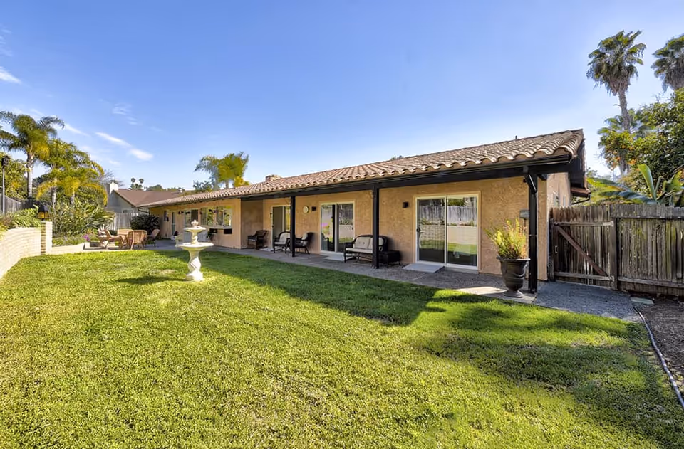 Backyard view of a single-story stucco building with a covered patio, sliding glass doors, green lawn and a small fountain.