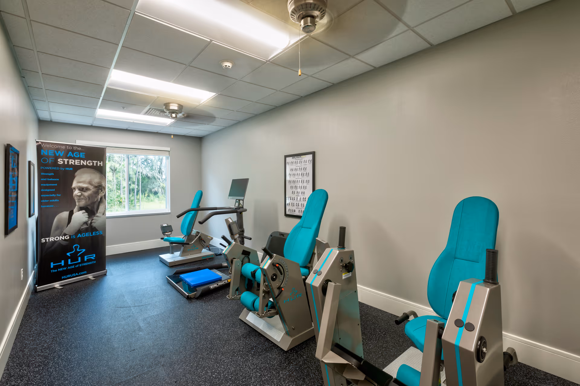 A small fitness room with exercise equipment including two seated strength training machines and a recumbent bike. The room has a window with a view of greenery outside, fluorescent ceiling lights, and a poster on the left side promoting strength training for seniors.