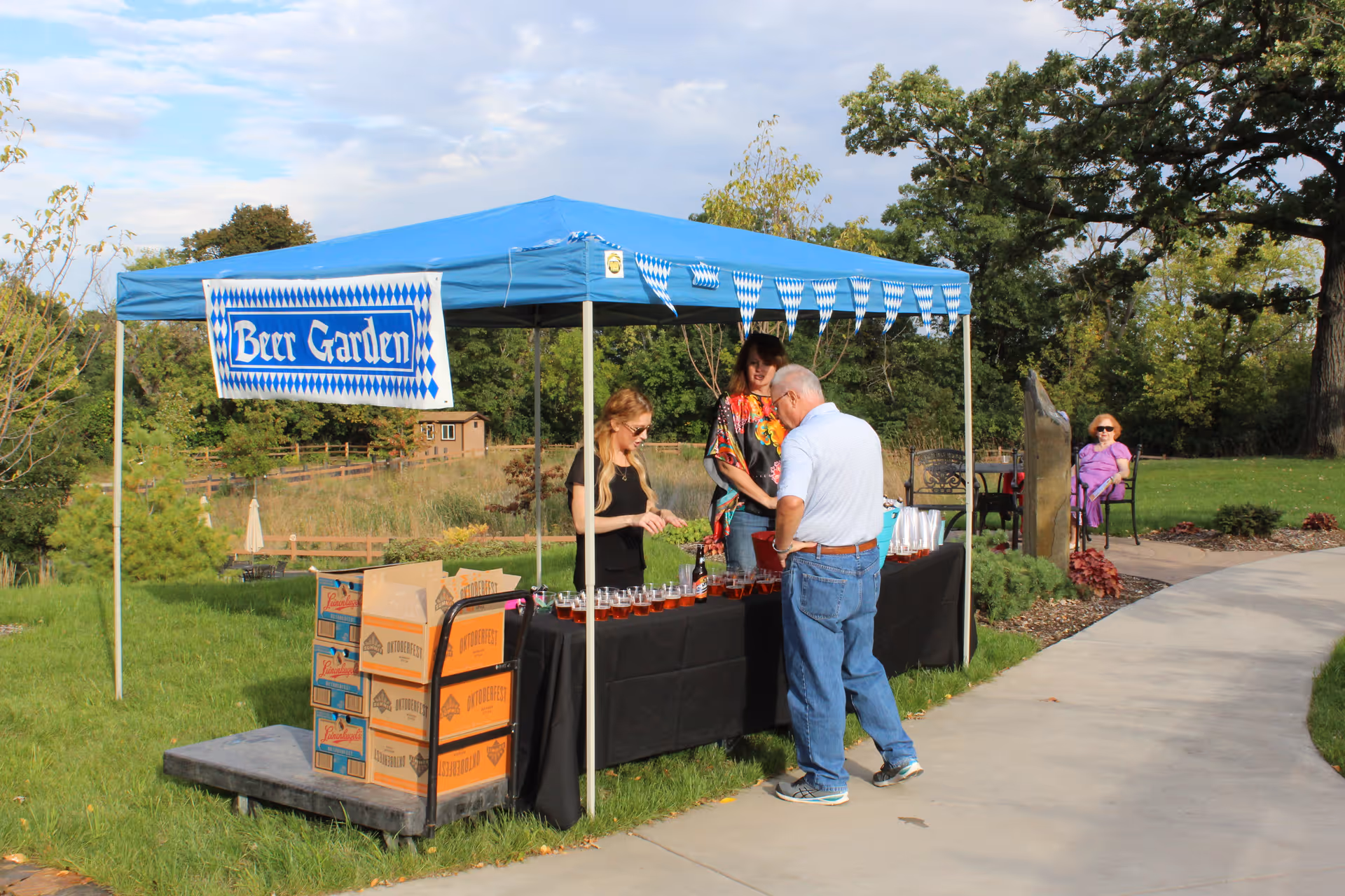An outdoor beer garden setup with a blue canopy tent and a 'Beer Garden' sign. Two women are serving drinks to an elderly man at a table covered with a black tablecloth. Several boxes of beverages are stacked on a dolly next to the table. In the background, a woman in purple is sitting on a bench along a paved pathway surrounded by green grass and trees.