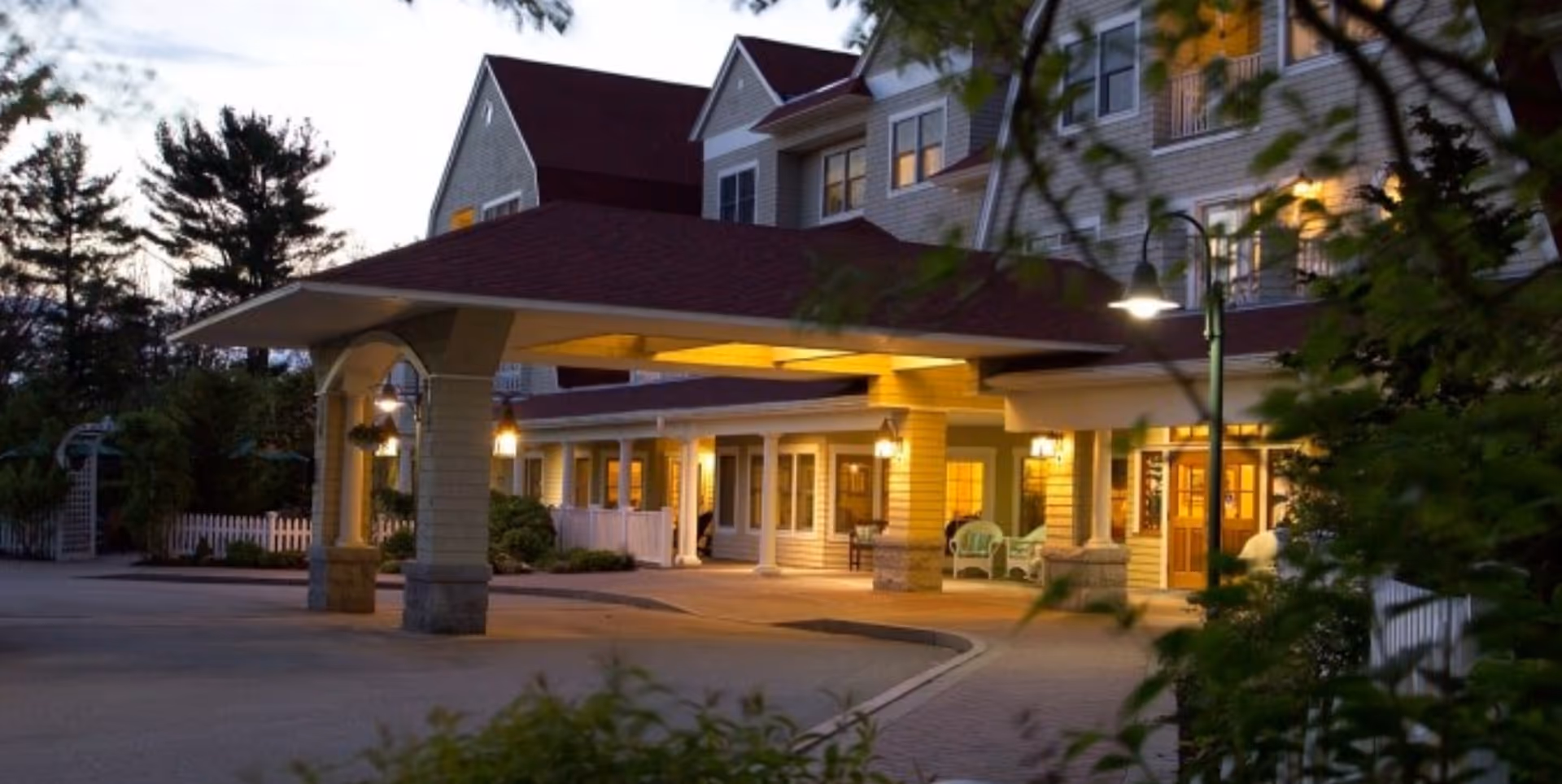 Front entrance of a multi-story senior living building with a covered porte-cochère lit at dusk.