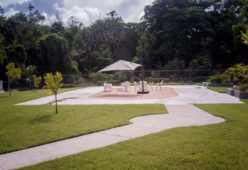 Outdoor seating area in a garden with a concrete pathway leading to a circular patio. The patio has a large umbrella providing shade over several white chairs and a table. The area is surrounded by green grass, small trees, and dense foliage in the background.