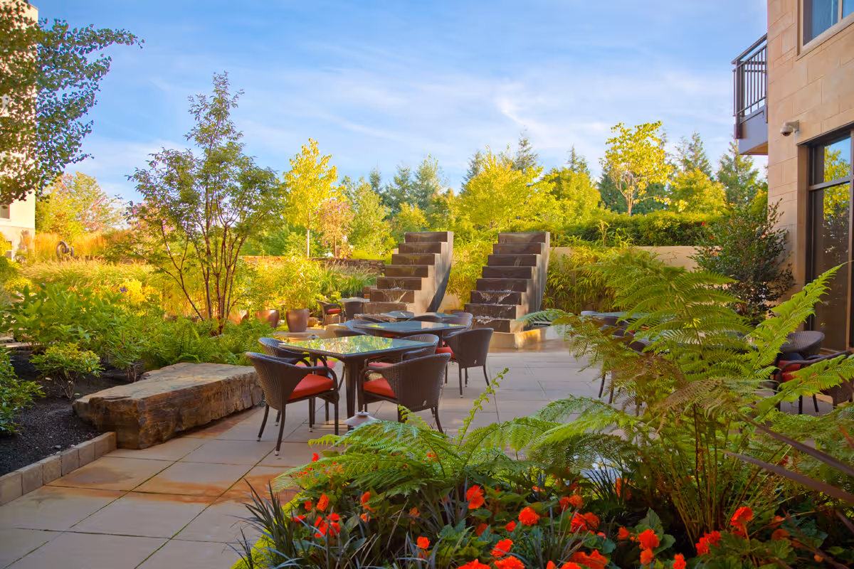 Outdoor patio area with glass-top tables and wicker chairs with red cushions surrounded by lush greenery, colorful flowers, and two modern water features with cascading water steps under a clear blue sky.