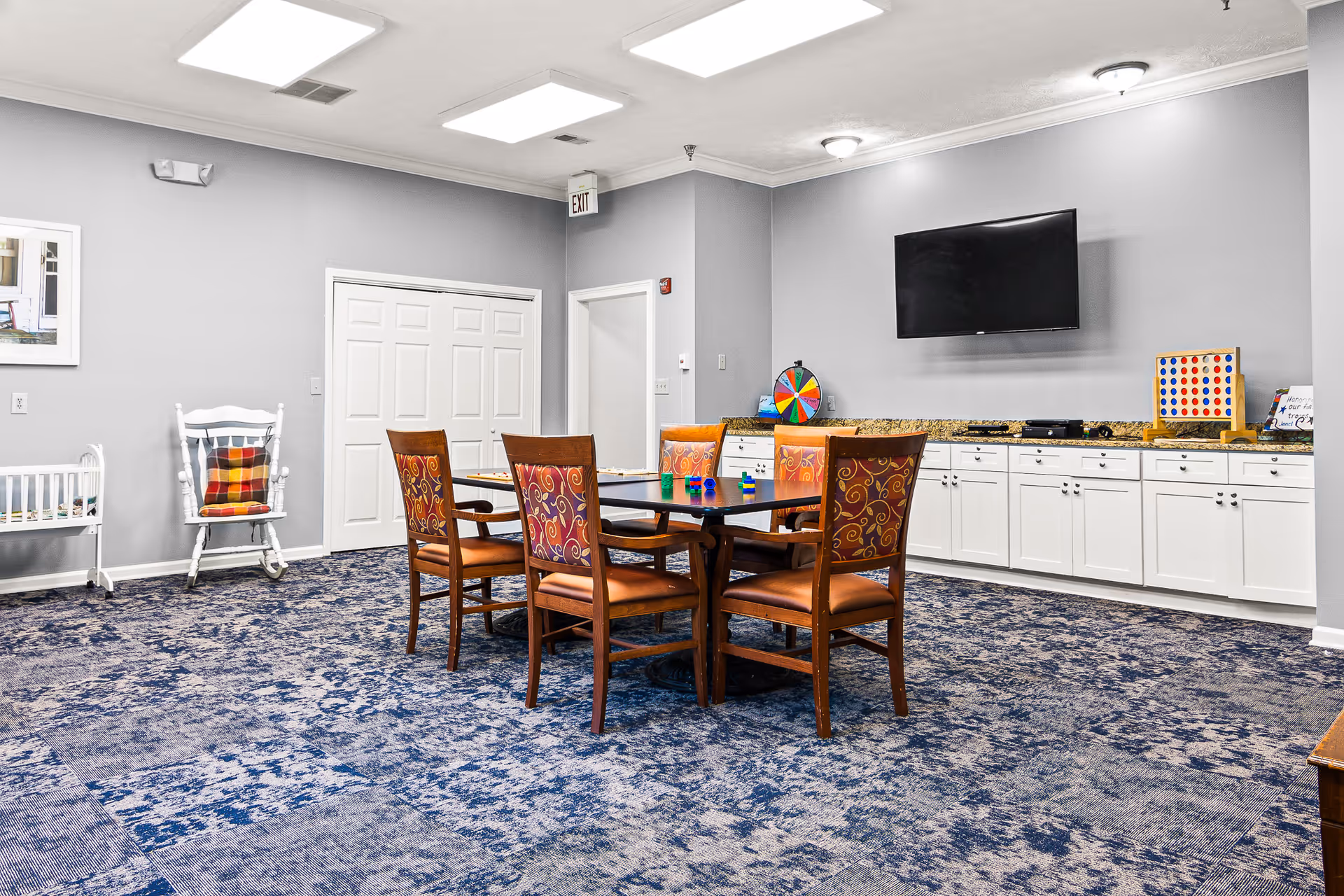 A well-lit activity room with a square table surrounded by six wooden chairs with patterned upholstery. The room has a blue patterned carpet, gray walls, and white cabinetry along one wall. On the countertop are a colorful spinning wheel, a Connect Four game, and some small toys. A flat-screen TV is mounted on the wall above the cabinets. There is a white rocking chair with a plaid cushion and a small white crib or bassinet against the opposite wall. An exit sign is visible above a door in the corner.