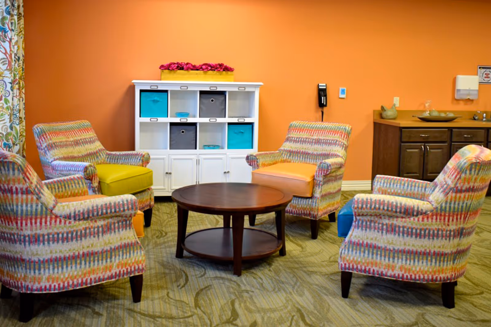 A cozy sitting area with four colorful patterned armchairs arranged around a round wooden coffee table. Behind the chairs is a white storage unit with cubbies containing blue and gray storage bins, topped with a yellow box filled with pink flowers. The walls are painted orange, and there is a wooden cabinet with a sink and decorative items on the right side.