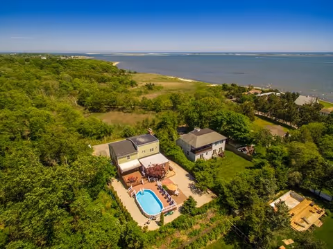 Aerial view of a residential property with a swimming pool, surrounded by trees and greenery, near a large body of water under a clear blue sky.