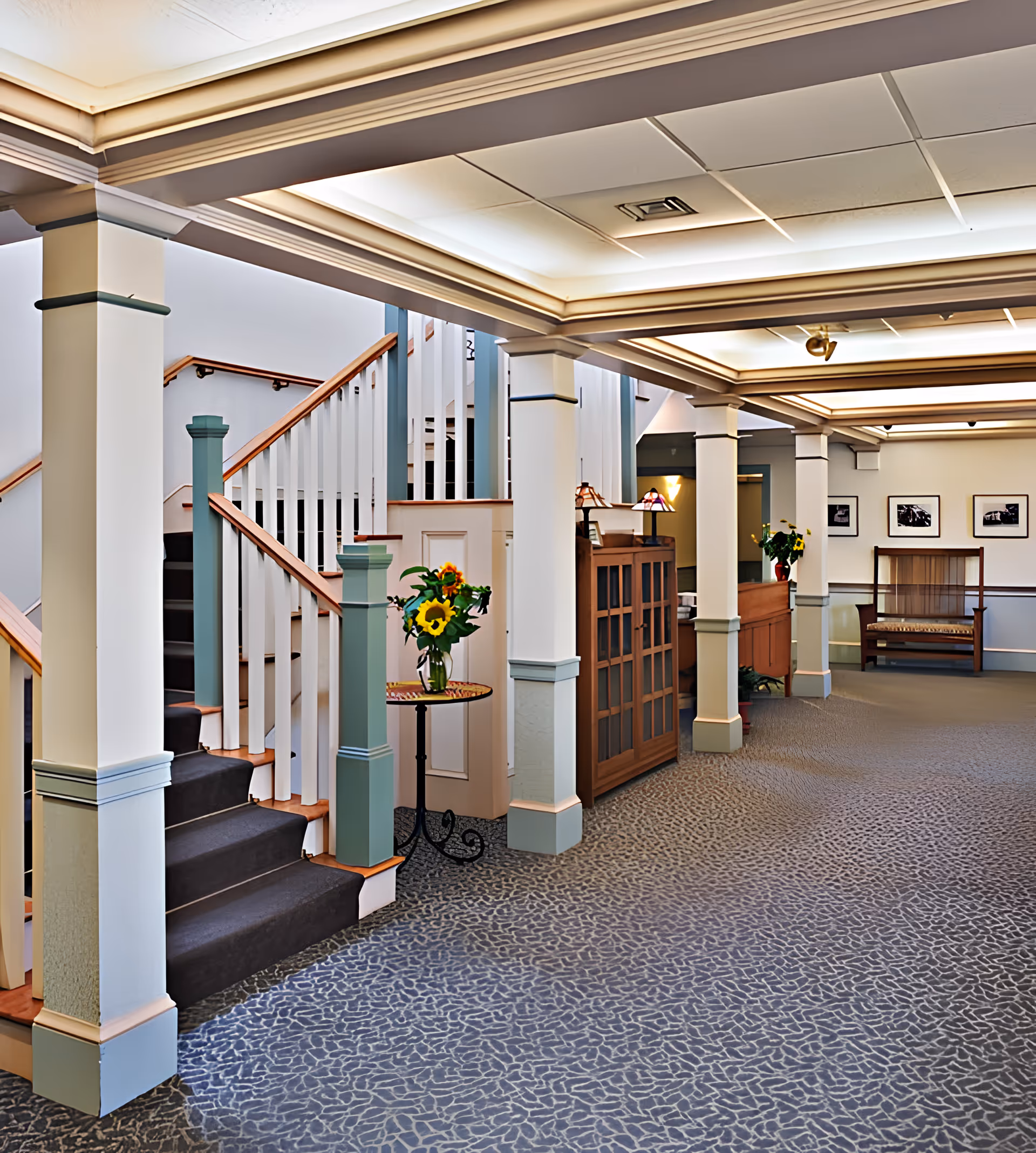 Well-lit interior lobby with a staircase, columns, seating and small tables with flower arrangements.