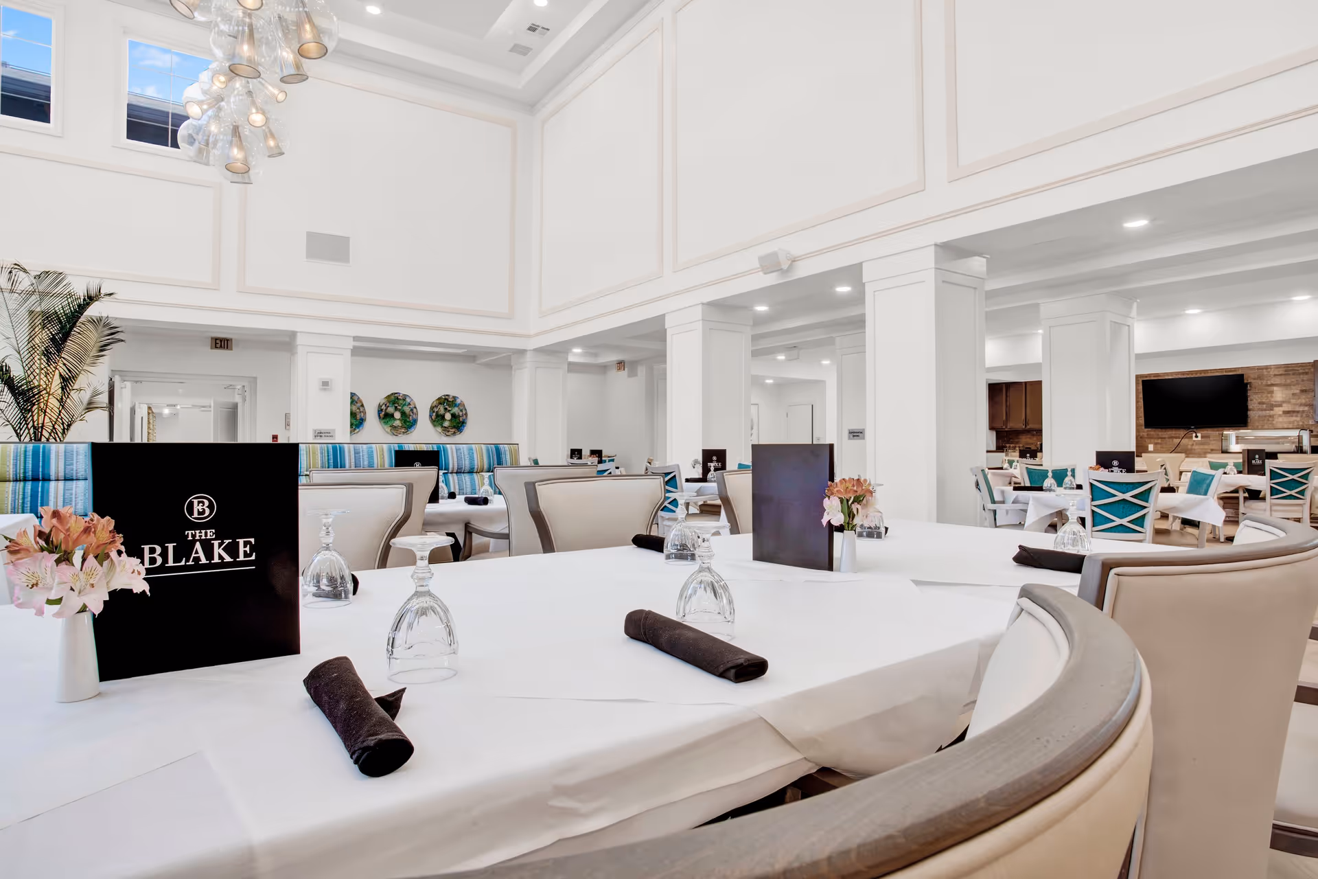 Bright and spacious dining room with white walls and high ceilings. Tables are set with white tablecloths, black napkins, upside-down wine glasses, and small flower arrangements. Comfortable chairs surround the tables, and a large black menu with 'The Blake' logo is prominently displayed on one table. The room features modern light fixtures, decorative wall plates, and a TV mounted on a wooden accent wall in the background.
