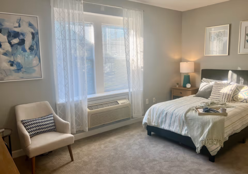 Sunlit bedroom with a bed, bedside table and lamp, an armchair, and a large window with sheer curtains.
