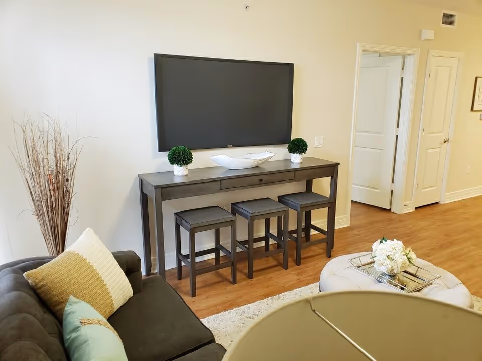 A cozy living room with a dark gray couch adorned with decorative pillows, a wooden console table with three stools underneath, two small potted plants, and a decorative bowl on top. A large flat-screen TV is mounted on the wall above the console table. There is a round ottoman with a tray and flowers in the foreground, and an open doorway leading to another room in the background. The floor is wooden, and the walls are painted light beige.