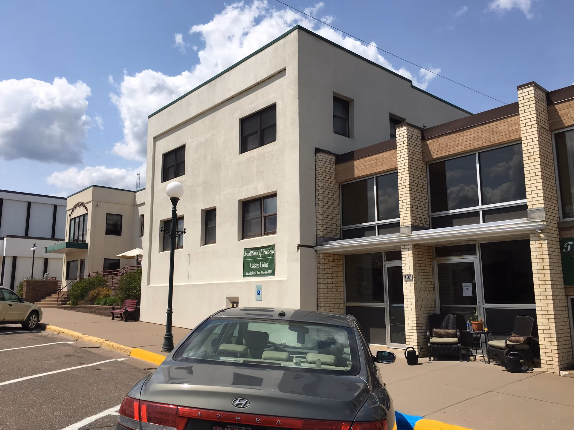 Exterior view of Traditions of Frederic assisted living facility showing a beige and brick building with multiple windows, a sidewalk with a bench, a street lamp, and parked cars under a partly cloudy sky.