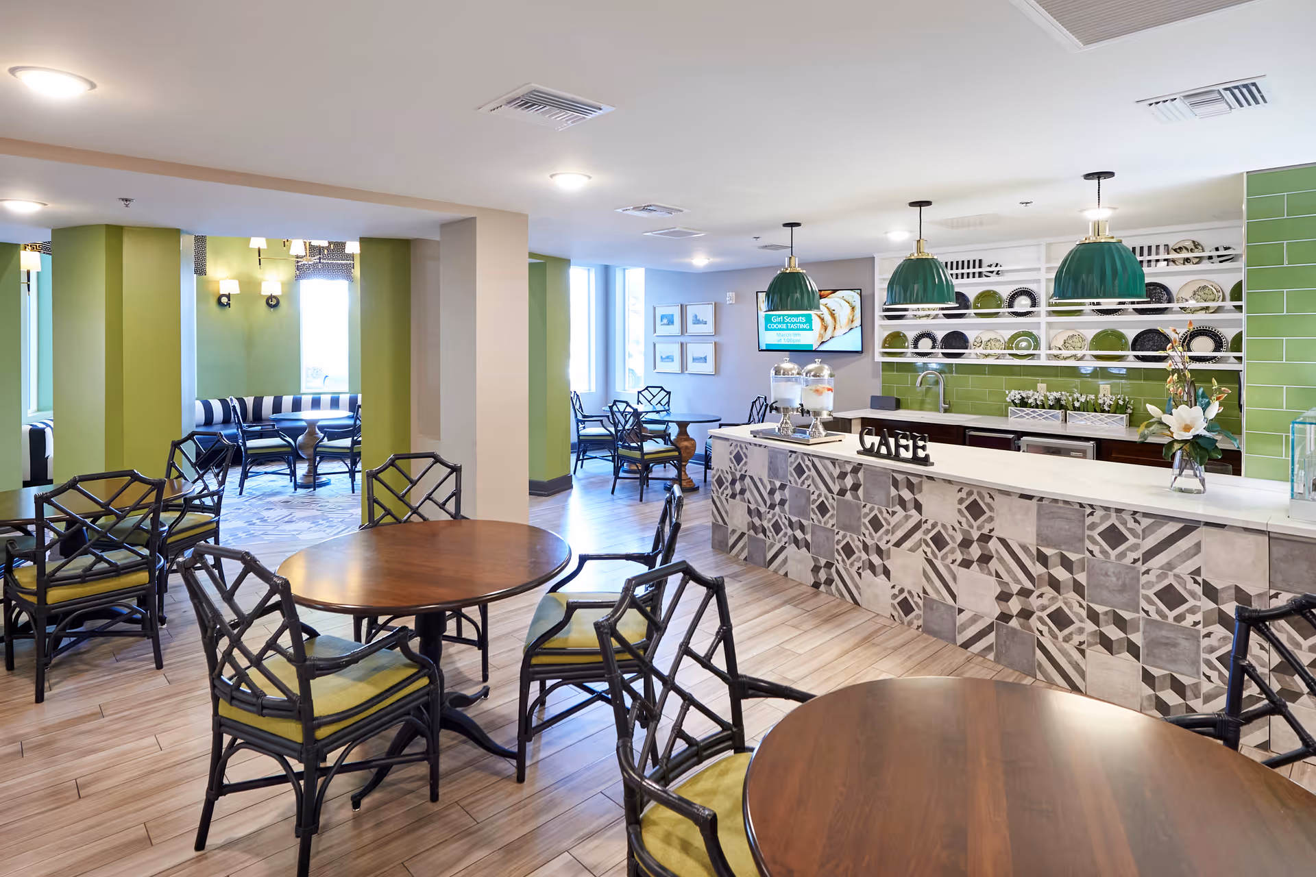 A bright and modern cafe area in a senior living facility with round wooden tables and black chairs with green cushions. The cafe counter features a patterned tile front and a white countertop with decorative items and a sign that reads 'CAFE'. Behind the counter, there are green tiled walls and shelves displaying plates. Green pendant lights hang above the counter. In the background, there is a seating area with striped cushioned benches and additional chairs near windows letting in natural light.