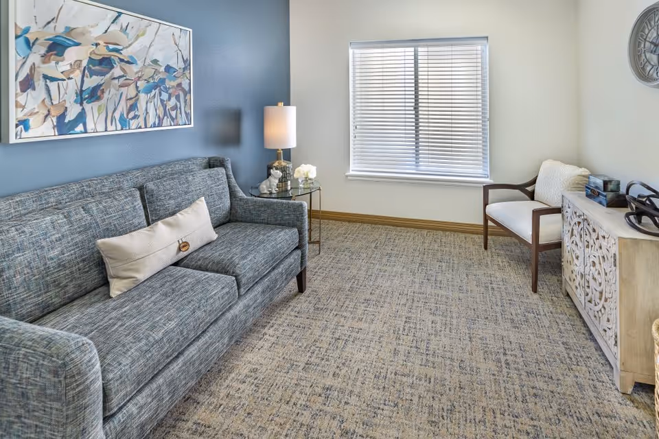 A cozy living room with a blue-gray sofa adorned with a beige pillow, a side table with a lamp and decorative items, a window with blinds, a wooden chair with a cushion, a decorative wooden cabinet, and a large wall clock. The walls are painted in blue and white, and the floor is carpeted.
