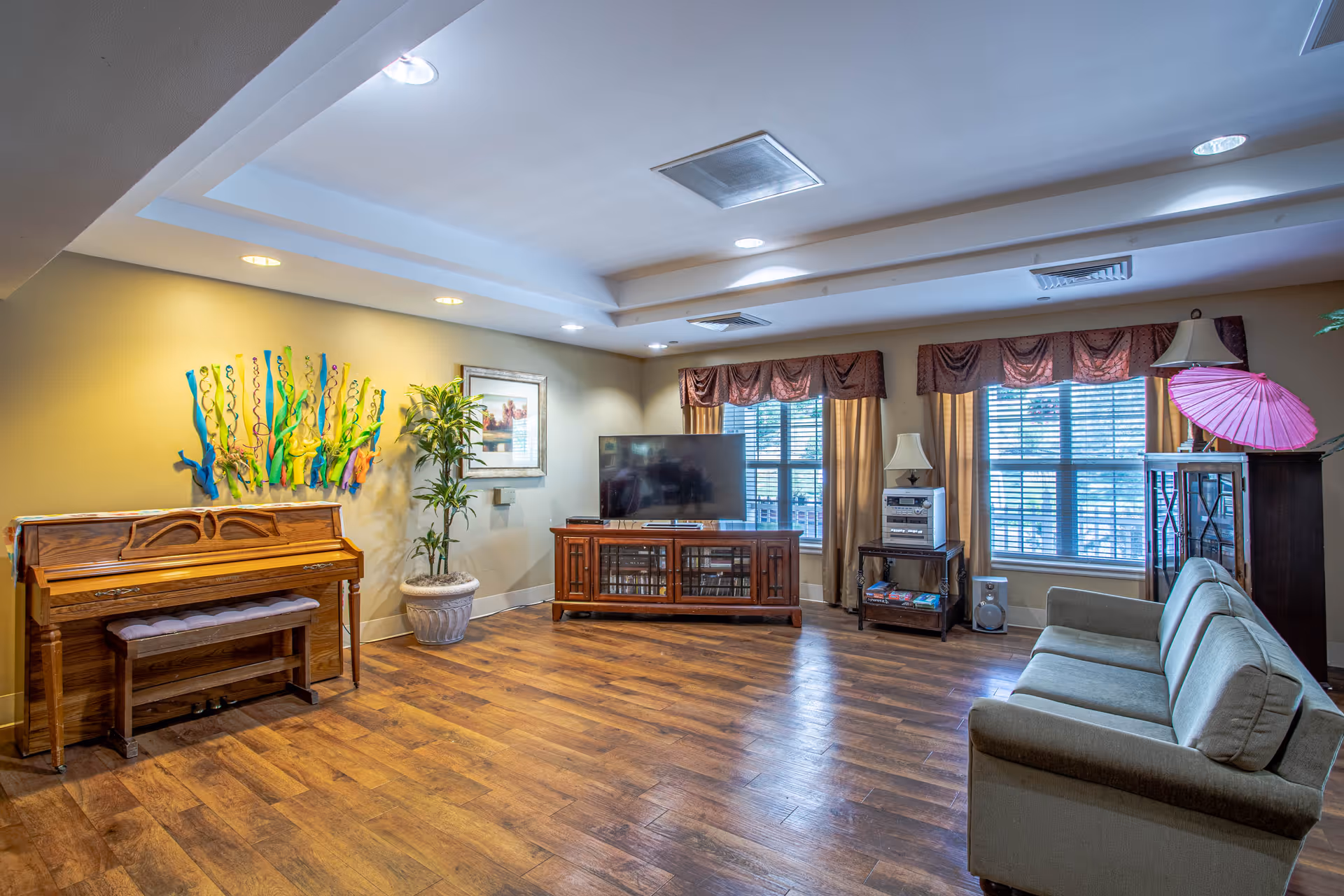 A spacious living room with wooden flooring, a piano with a bench on the left, a flat-screen TV on a wooden stand in the center, two large windows with curtains, a gray sofa on the right, and decorative items including a plant, a lamp, and a pink parasol.