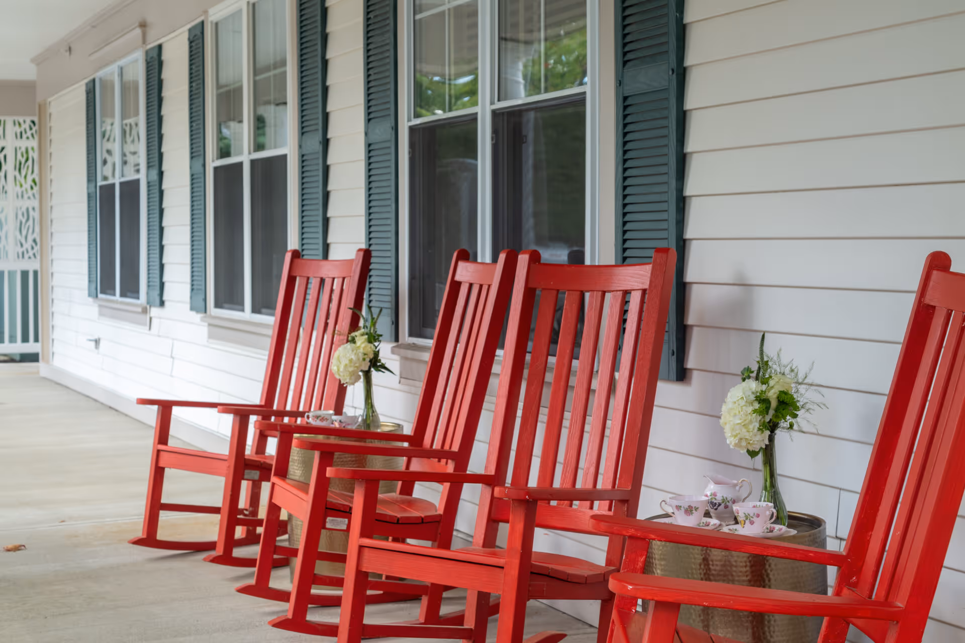 A porch area with four bright red wooden rocking chairs lined up against a white exterior wall with green shutters. Two small round tables between the chairs hold vases with white flowers and a tea set.