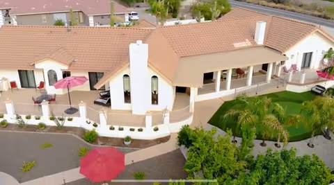 Aerial view of a single-story assisted living facility with a terracotta tiled roof, outdoor patio areas with red umbrellas, and landscaped grounds including palm trees and a putting green.