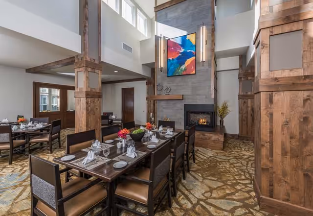 A spacious dining room with a long wooden table set for a meal, including plates, glasses, napkins, and floral centerpieces. The room features high ceilings with large windows, wooden beams and columns, a stone fireplace with a colorful abstract painting above it, and carpeted flooring with a nature-inspired pattern.