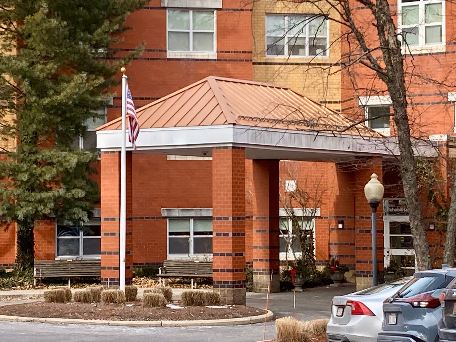 Entrance area of Spaulding Nursing & Therapy Center showing a covered drop-off with a copper-colored roof supported by brick pillars. There is an American flag on a flagpole in front, benches on either side, some trees, and parked cars nearby.