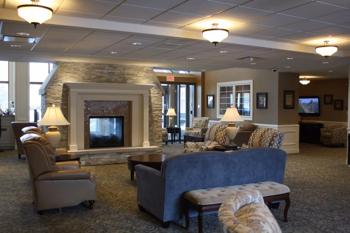 A cozy senior living facility common area with a stone fireplace as the focal point. The room features comfortable seating including leather armchairs, patterned armchairs, a blue sofa, and a cushioned bench. Several table lamps provide warm lighting, and a television is visible in the background. The space has carpeted floors and a drop ceiling with recessed lighting.