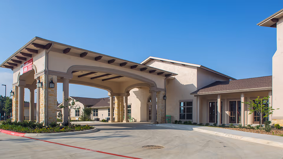 Exterior view of the entrance to Franklin Park Boerne senior living facility featuring a covered drop-off area with stone columns and beige walls under a clear blue sky.