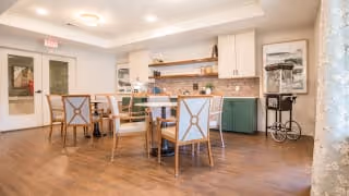 Well-lit dining room with round tables and chairs facing a small kitchenette with cabinets, shelving, and artwork.