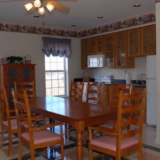 A dining area with a wooden table and six matching wooden chairs with pink cushions. The room has a ceiling fan with lights, a window with a blue valance, and a kitchen area in the background featuring wooden cabinets, a microwave, stove, and refrigerator. There is also a wooden cabinet with plants on top near the window.