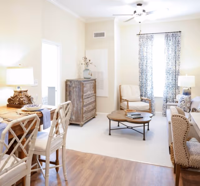 Bright open living room with armchairs, a round coffee table, patterned curtains, and a dining table with chairs in the foreground.