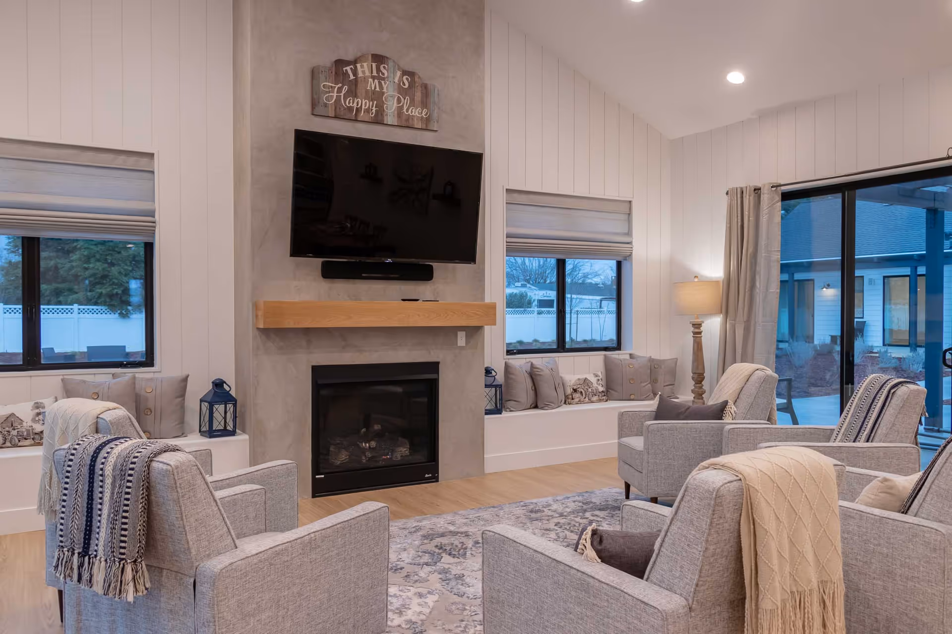 A cozy living room with four gray armchairs arranged around a fireplace with a mounted flat-screen TV above it. The room features light wood flooring, a patterned area rug, and large windows with light-colored blinds. There are decorative pillows on built-in window seats and a floor lamp in the corner. A sign above the TV reads 'This is my Happy Place.'