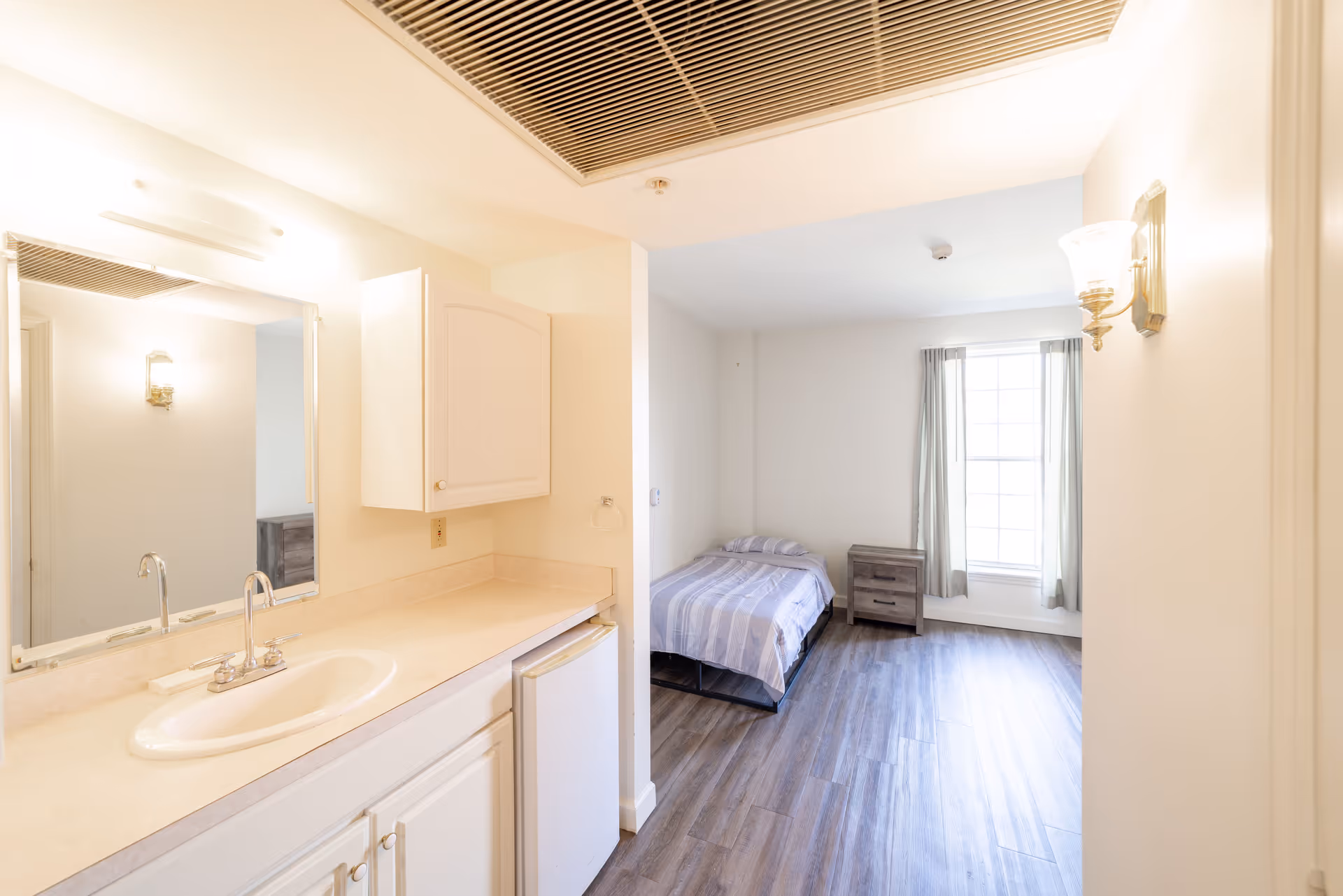 A bright room with a small bed covered in a striped blanket, a wooden nightstand, and a large window with curtains. Adjacent to the room is a vanity area with a sink, countertop, mirror, and a small refrigerator underneath the counter. The floor is wood-style laminate and the walls are painted white.