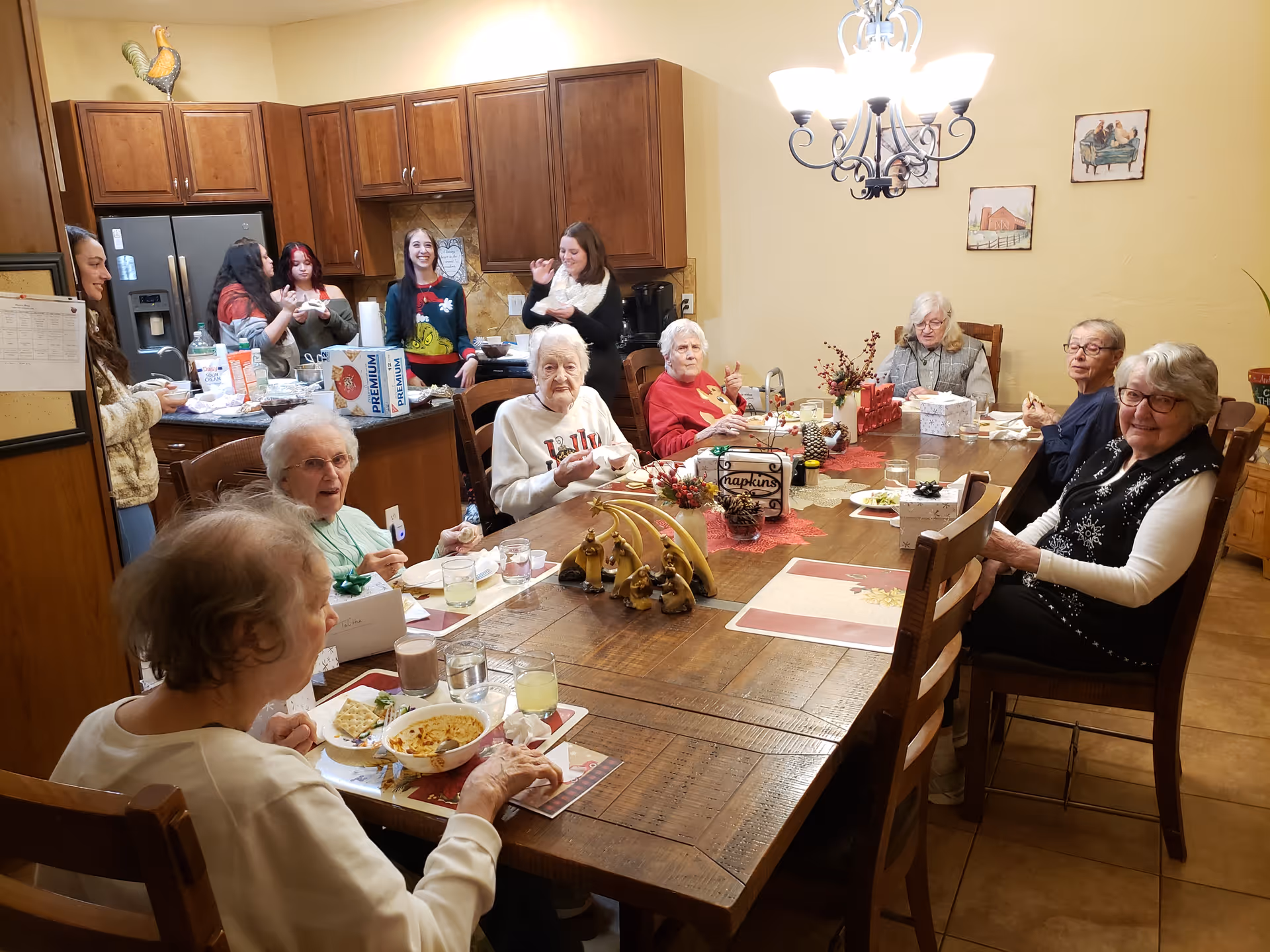 Several elderly residents and caregivers seated and standing around a large dining table in a communal kitchen/dining area.