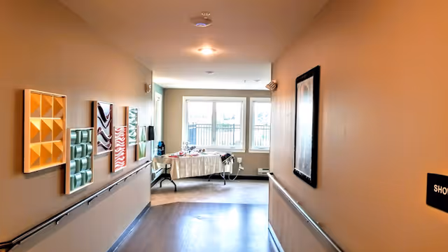 A well-lit hallway with handrails and wall art leading to a windowed common area with a table at the end.