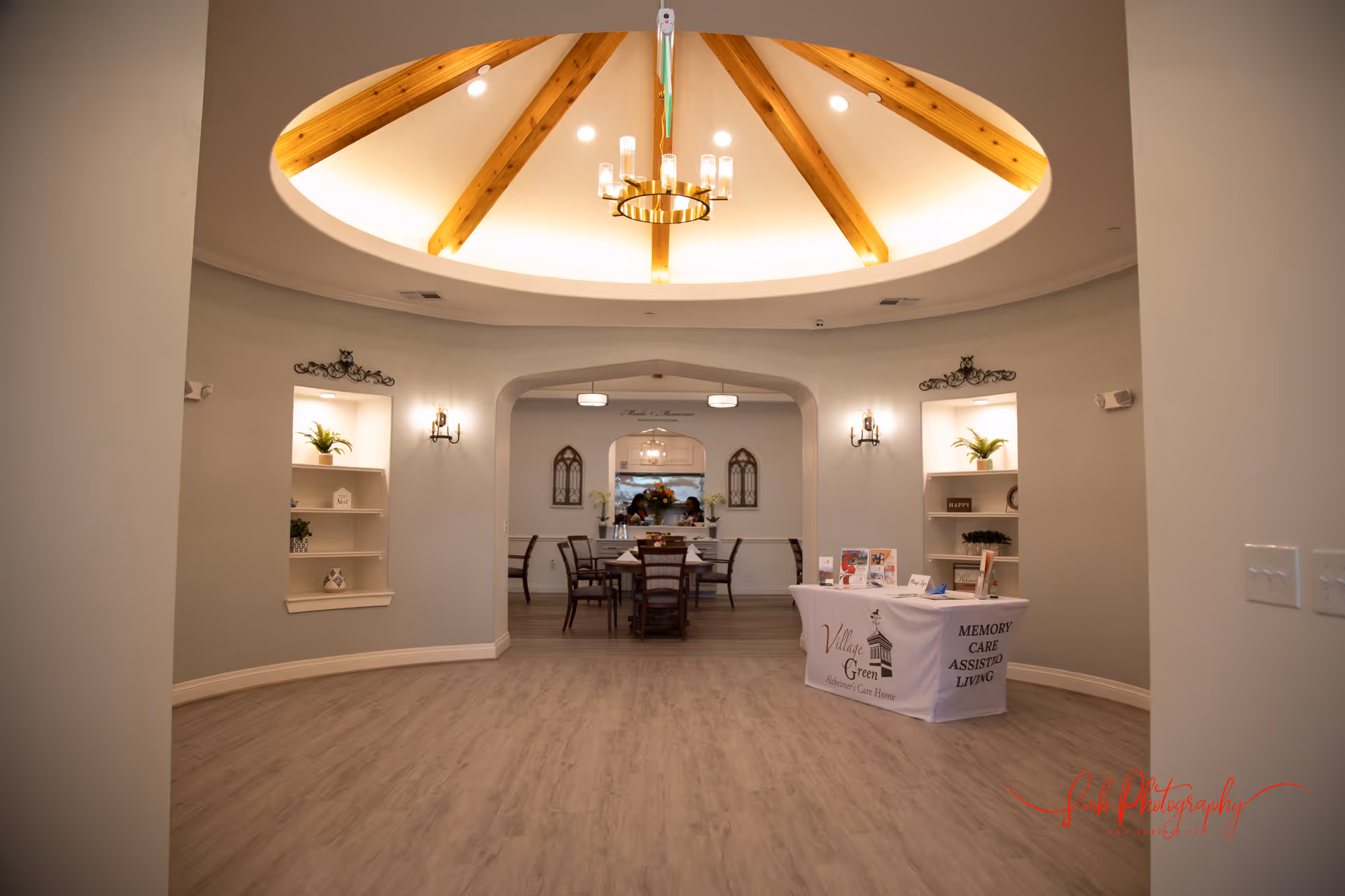 Interior view of a senior living facility with a circular ceiling featuring wooden beams and a chandelier. The room has light-colored walls and wood flooring. There are built-in shelves with plants and decorative items on either side of an arched doorway leading to a dining area with a table and chairs. A table covered with a cloth displaying 'Village Green Memory Care Assisted Living' is positioned on the right side near the entrance.