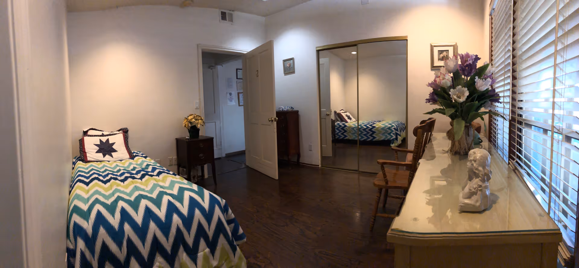 A small bedroom with a single bed covered in a blue, green, and white chevron-patterned blanket and a pillow with a star design. There is a wooden nightstand with a small plant next to the bed. The room has wooden floors, a door slightly ajar, and a mirrored closet door reflecting the bed. A wooden chair and a long dresser with a vase of flowers and a small statue are positioned along the window with blinds.
