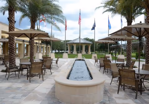 Outdoor courtyard with palm trees, shaded patio tables, a central water fountain, and a gazebo with flags in the background.