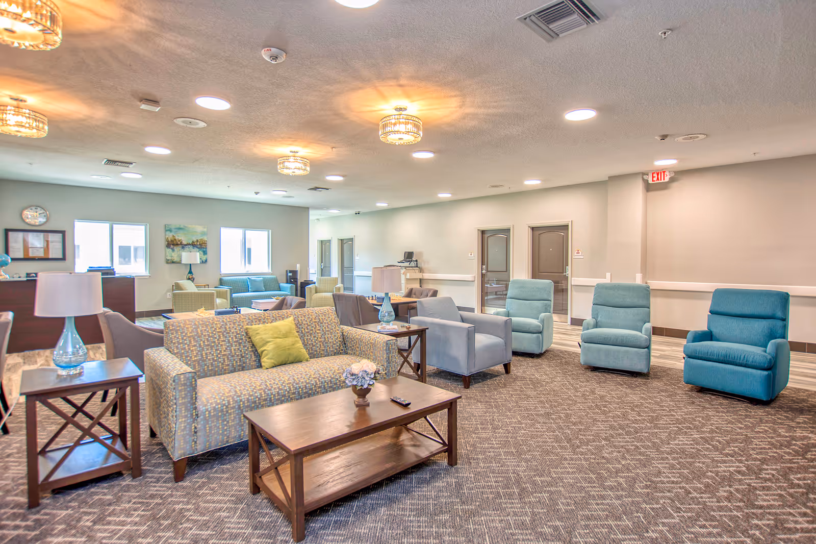 A spacious and well-lit common area in an assisted living facility featuring multiple seating options including patterned sofas, blue armchairs, and side tables with lamps. The room has a carpeted floor, neutral-colored walls, ceiling lights, and windows allowing natural light. There are doors along the back wall and an exit sign above one door.