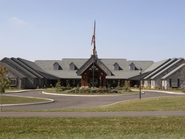 Front exterior view of Heritage Senior Living of Marysville, a large single-story building with a peaked roof, stone facade, and an American flag flying on a flagpole in front. The driveway curves around a landscaped circular island with shrubs and flowers.