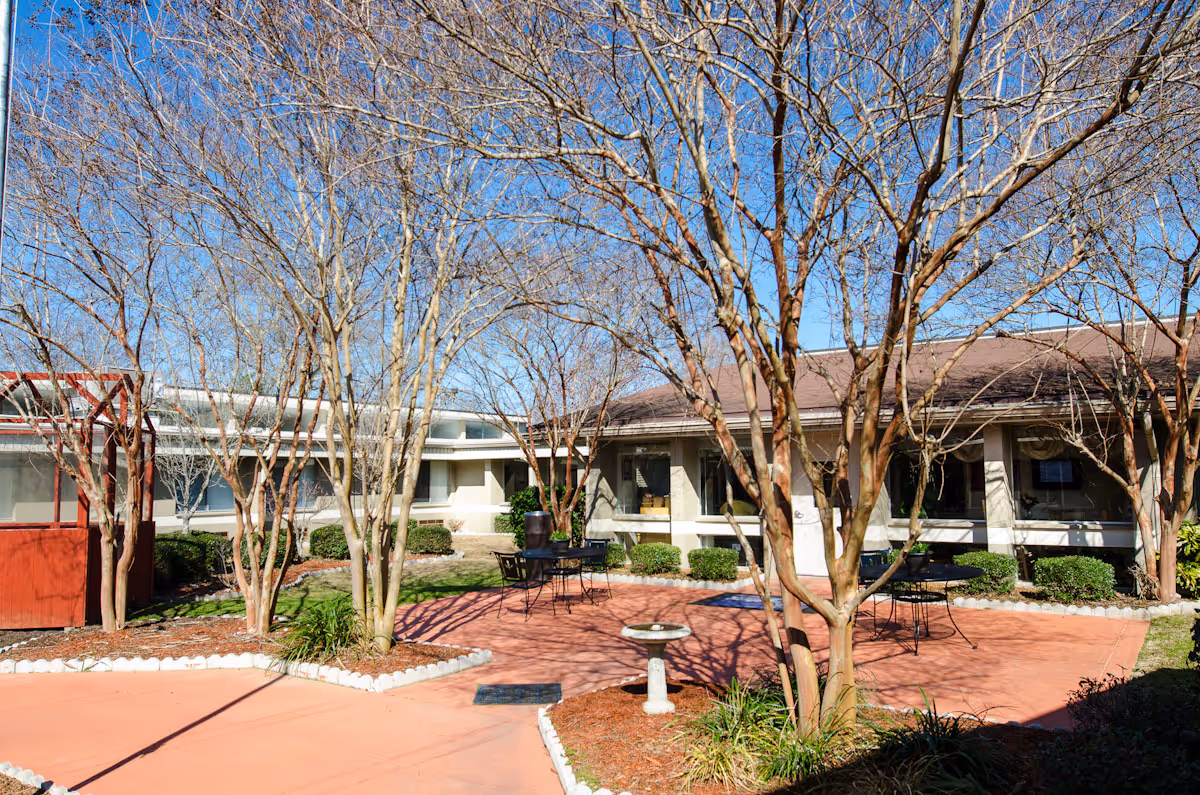 Outdoor courtyard area at Graceville Health Center with leafless trees, a birdbath, black metal tables and chairs, and a building with large windows in the background under a clear blue sky.