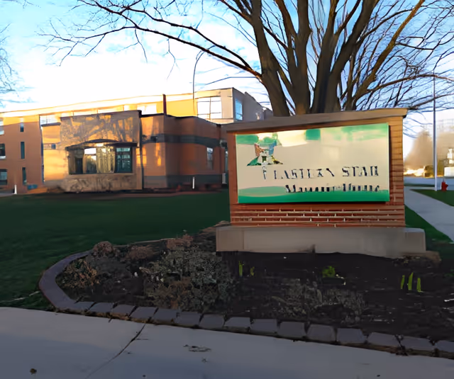 Outdoor view of the Eastern Star Masonic Home building with a large tree and a sign in front displaying the facility's name. The building is made of brick and has multiple windows. There is a sidewalk and a grassy area surrounding the sign and building.