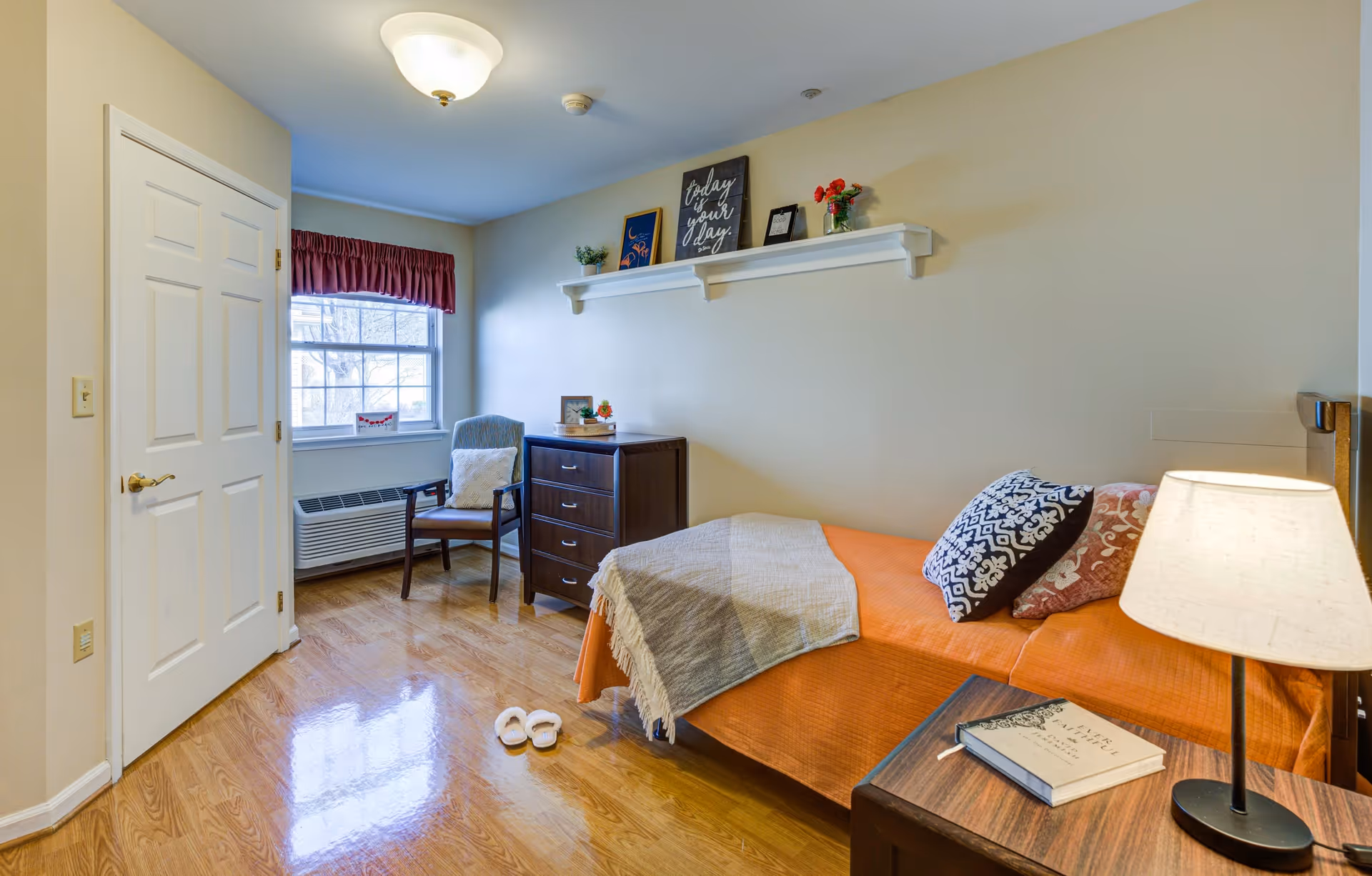 A cozy bedroom with a single bed covered in an orange bedspread and decorative pillows. A beige throw blanket is draped over the bed. Next to the bed is a wooden nightstand with a lamp and a book. Across from the bed is a wooden dresser with a chair beside it. Above the dresser is a white wall shelf holding framed pictures, a small plant, and a vase with red flowers. A window with a red valance allows natural light into the room, and a pair of white slippers is placed on the wooden floor near the bed.
