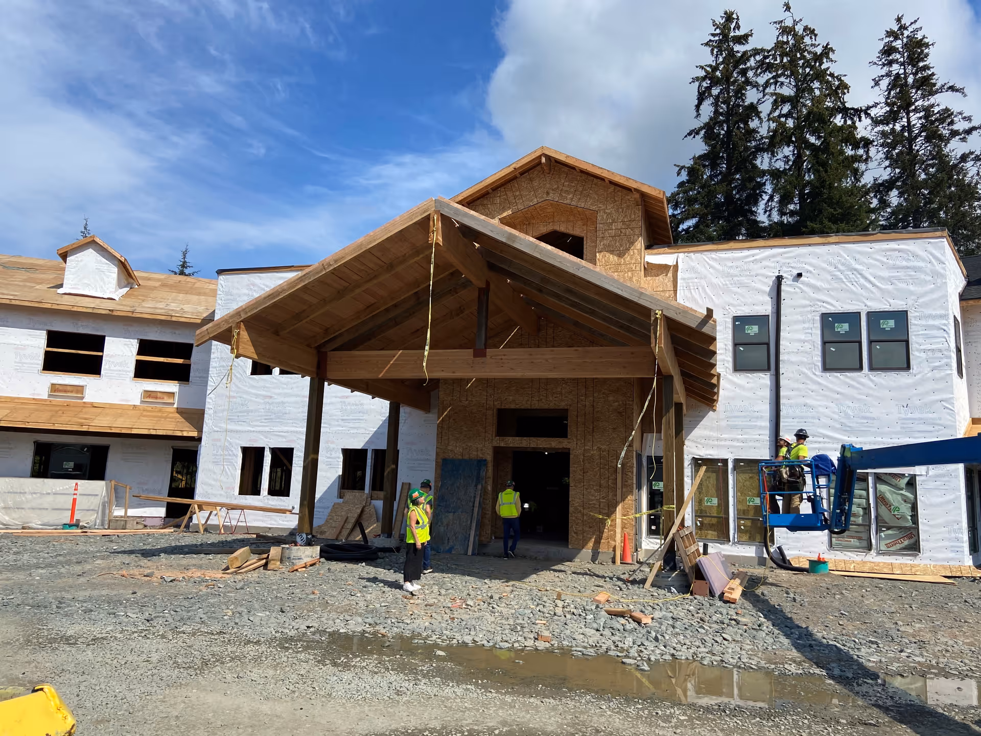 Construction site of a large building with wooden framing and white weatherproof sheeting. Several workers in yellow safety vests and helmets are present, some standing on the ground and others on a blue lift. The building appears to be in the early stages of construction with exposed wood and unfinished windows. Tall trees and a partly cloudy blue sky are visible in the background.