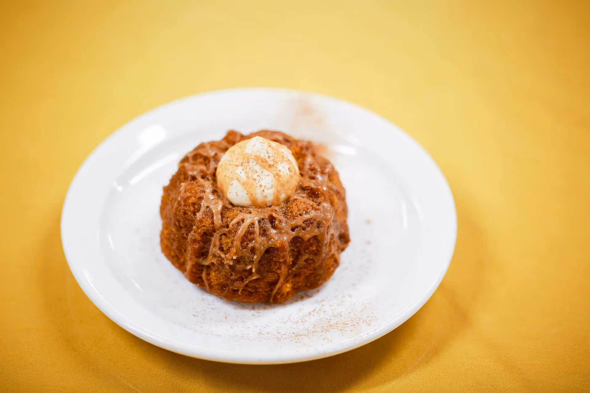 A close-up of a small bundt cake on a white plate, topped with a dollop of cream and sprinkled with cinnamon, placed on a yellow tablecloth.