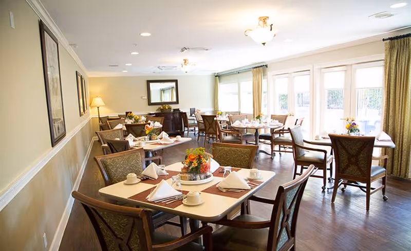 Bright dining room with multiple set tables and chairs, floral centerpieces, and large windows letting in natural light.