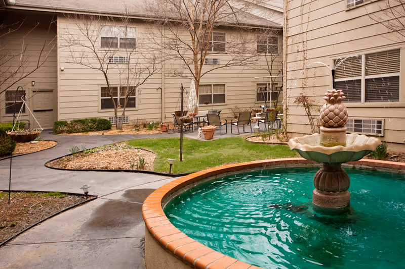 Outdoor courtyard area of Aberdeen Heights Assisted Living featuring a circular water fountain with a pineapple design, surrounded by a paved walkway, small grassy patches, leafless trees, and outdoor seating with tables and chairs near the beige building walls.
