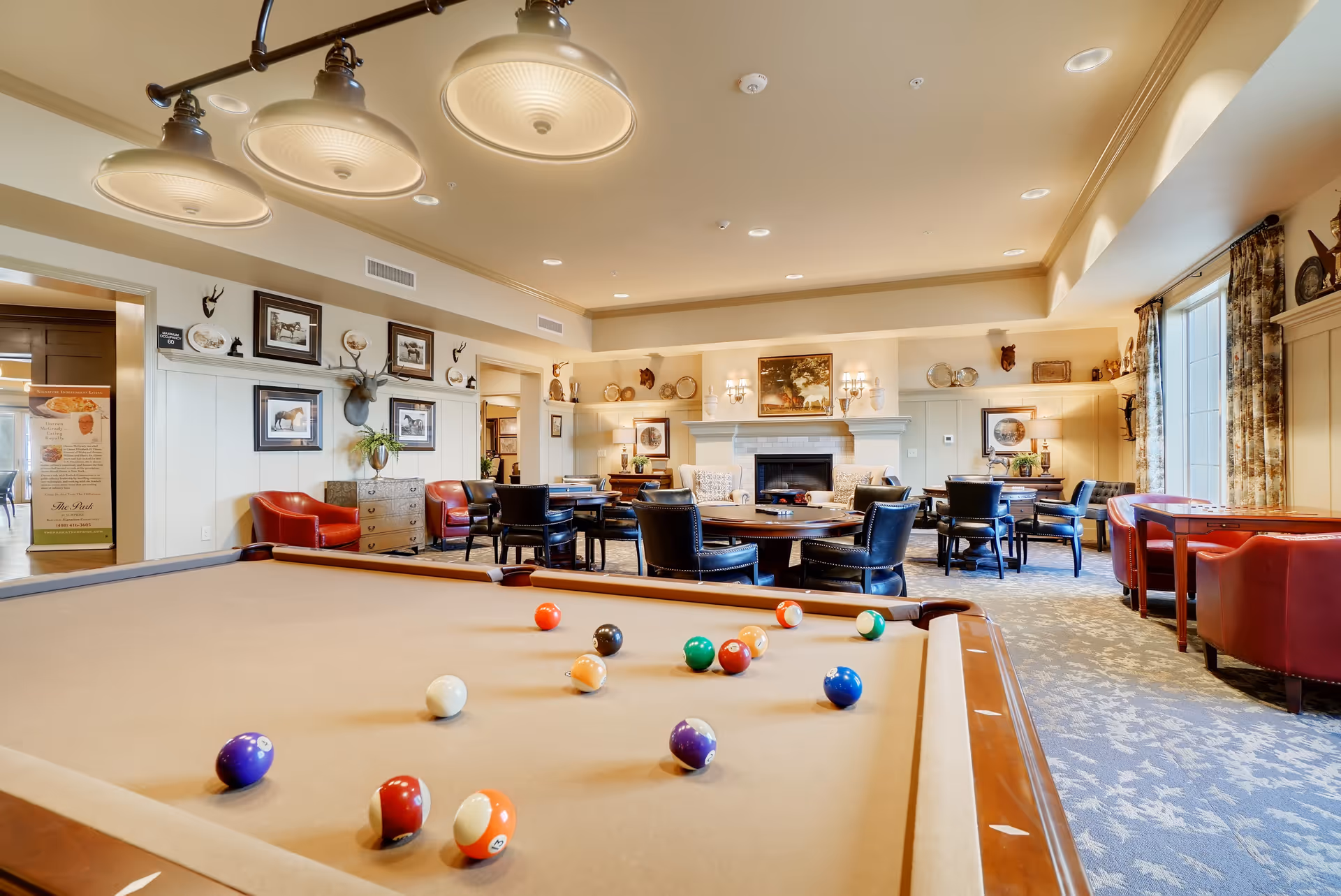 A spacious and well-lit common area in a senior living community featuring a pool table with billiard balls scattered on it, multiple round tables with black chairs, red armchairs, a fireplace with artwork above it, and large windows with patterned curtains allowing natural light to fill the room.