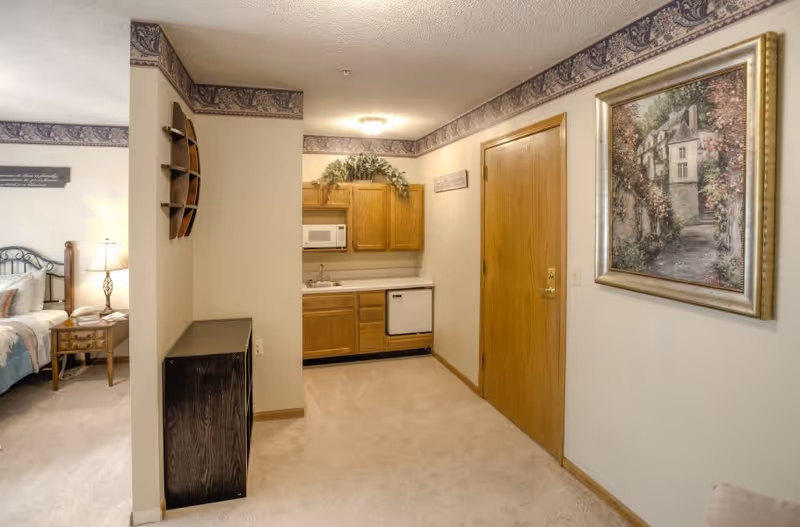 Interior view of a senior living facility room showing a small kitchenette with wooden cabinets, a microwave, a sink, and a dishwasher. To the left, part of a bedroom with a bed, nightstand, and lamp is visible. The walls have a decorative border near the ceiling, and a framed painting hangs on the right wall next to a wooden door.
