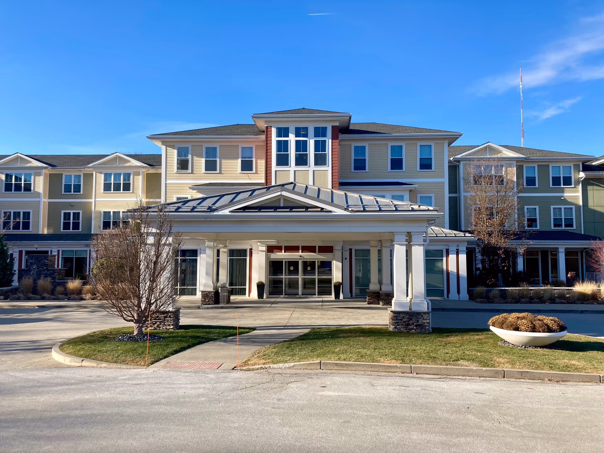 Front exterior view of a multi-story senior living facility building with a covered entrance, large windows, and a clear blue sky in the background. There is a driveway and landscaped areas with grass, trees, and a large planter in front.